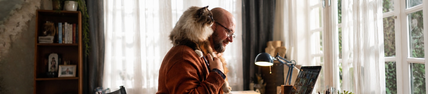 Man sitting at a computer smiling while a fluffy cat perches on his shoulder