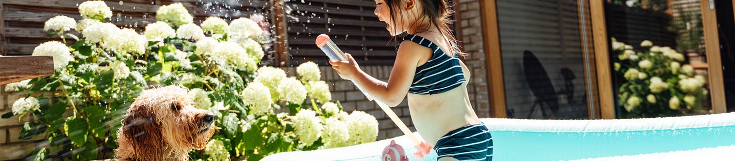 A little girl in a swimsuit stands in a turquoise paddling pool and sprays water from a toy while a shaggy light brown dog looks on